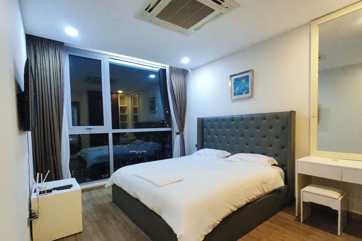 A bedroom with a large bed, a bedside table, a desk, a mirror, and a window with curtains.