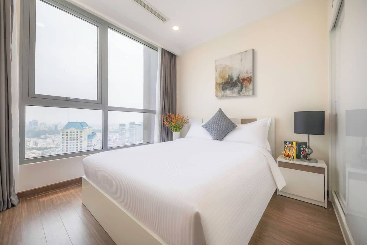 A hotel room with a large bed, a nightstand, a lamp, a painting, and a window with a city view.