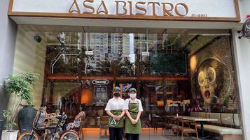 Exterior del ASA Bistro, con fachada de vidrio, se aprecian mesas y sillas en el interior a través de las ventanas, y un cartel que muestra el horario de atención.