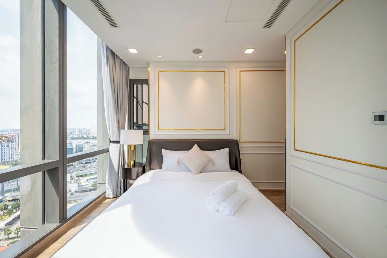 A hotel room with a large bed, a nightstand, a lamp, and a view of the city through large windows.