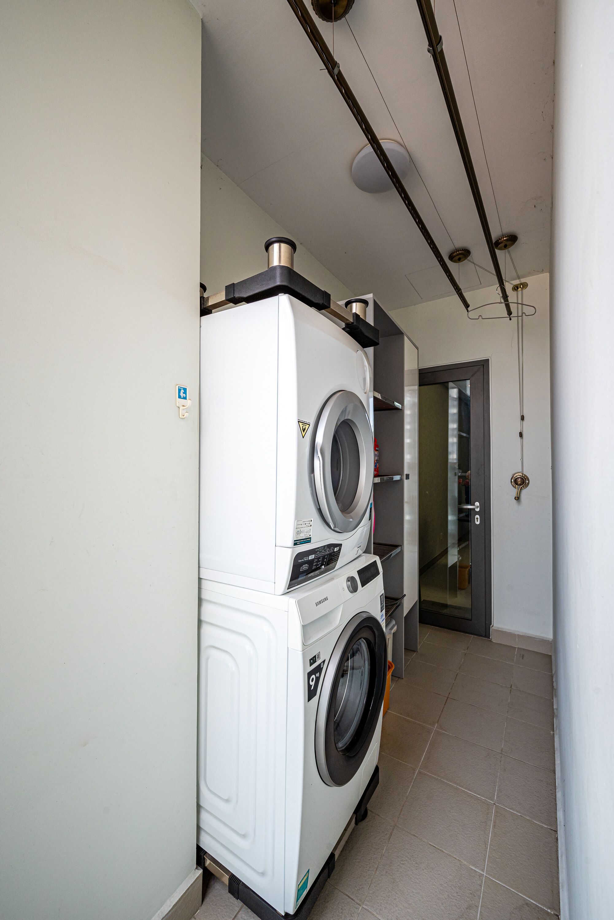 Une buanderie avec des machines à laver et des sèche-linge superposés, une porte et un plafond où les tuyaux sont apparents.