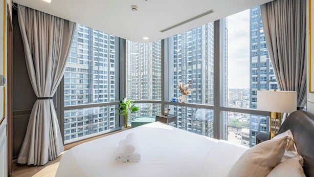 A modern hotel room with a large bed, a view of high-rise buildings, and a balcony with a plant. - Landmark 81 - Saigon Luxury Apartment (Ho Chi Minh City)