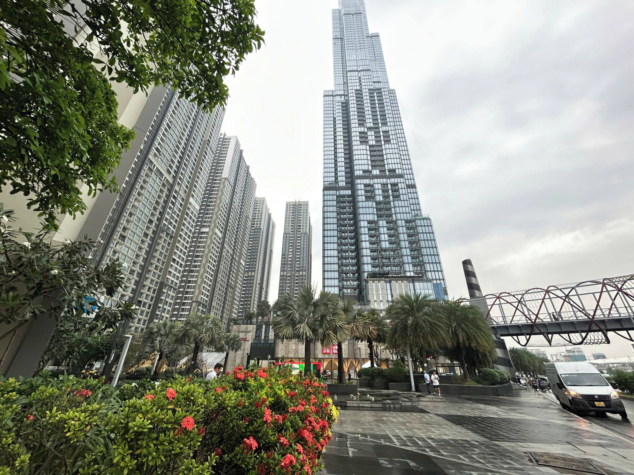 A cityscape with tall buildings, a bridge, and a pedestrian area with trees and flowers.
