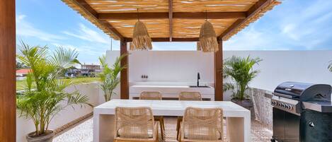 An outdoor patio with a wooden pergola, a white dining table with wicker chairs, a built-in grill, and potted plants.