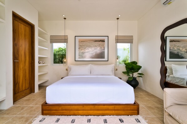 A bedroom with a wooden bed frame, white bedding, a large window with a view of the sea, and a mirror on the wall.