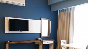 A hotel room with a blue wall, a flat-screen TV mounted above a wooden desk, a small bedside table, a yellow bench, a white chair, and a window with beige curtains. - EVLAND CİTY SUİT (Adana)