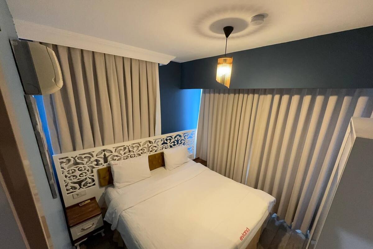A hotel room with a bed, curtains, a decorative headboard, a nightstand, and a ceiling light fixture.