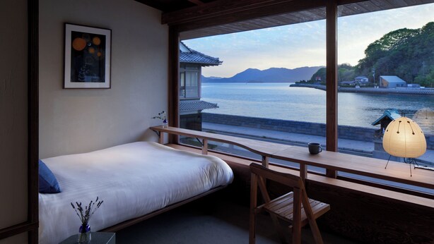 Una habitación con cama, escritorio, silla, ventana con vista a un cuerpo de agua y montañas, y un farol japonés tradicional.