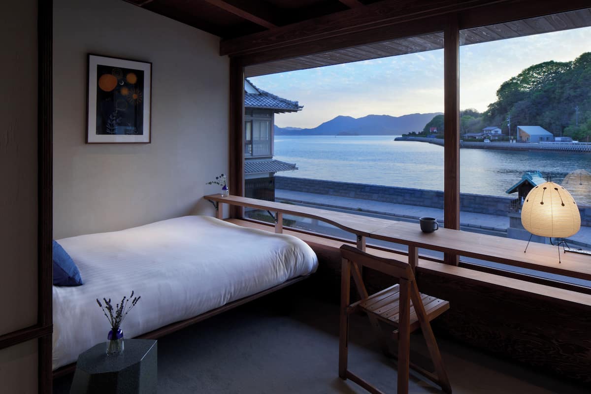 A room with a bed, a desk, a chair, a window with a view of a body of water and mountains, and a traditional Japanese lantern.