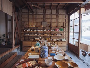 Ein rustikales Interieur mit Holzregalen, die verschiedene Gegenstände zeigen, einem Holztisch mit Schüsseln und Küchenutensilien sowie einem Blick durch ein großes Fenster.