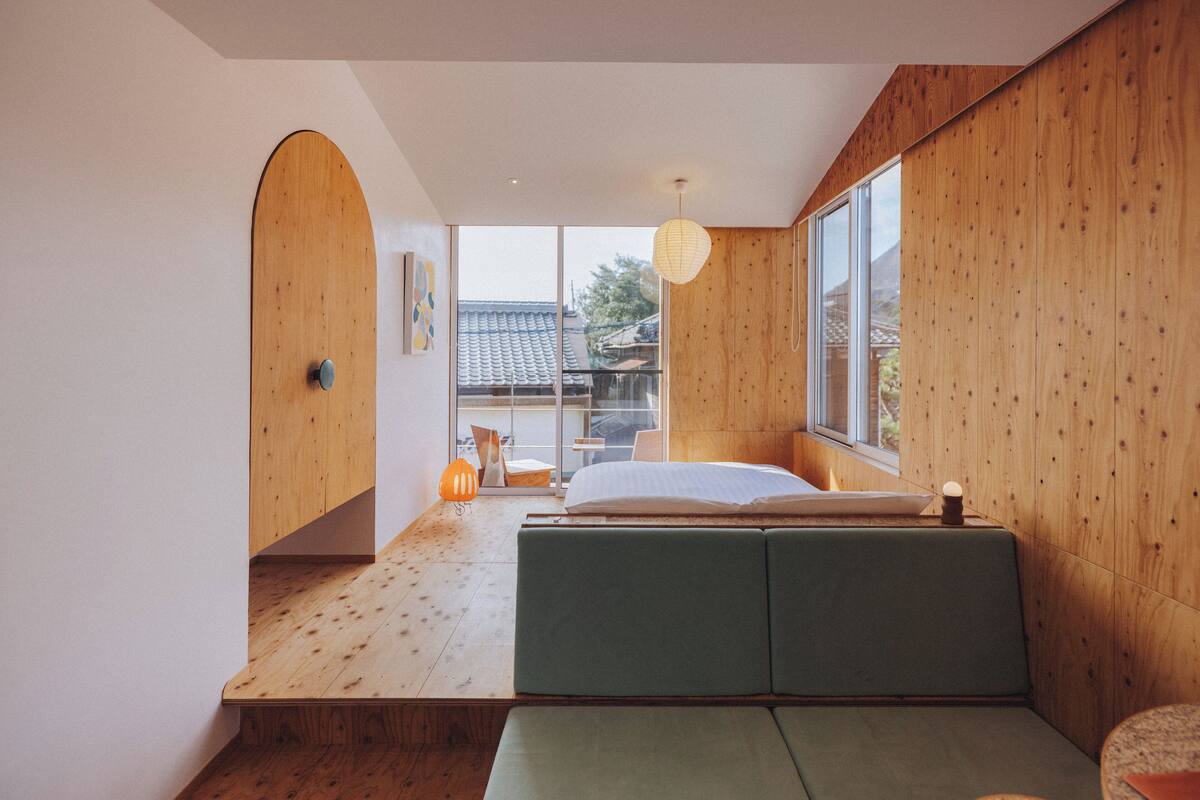A modern bedroom with a wooden floor, a green bed, and a large window offering a view of the outside.
