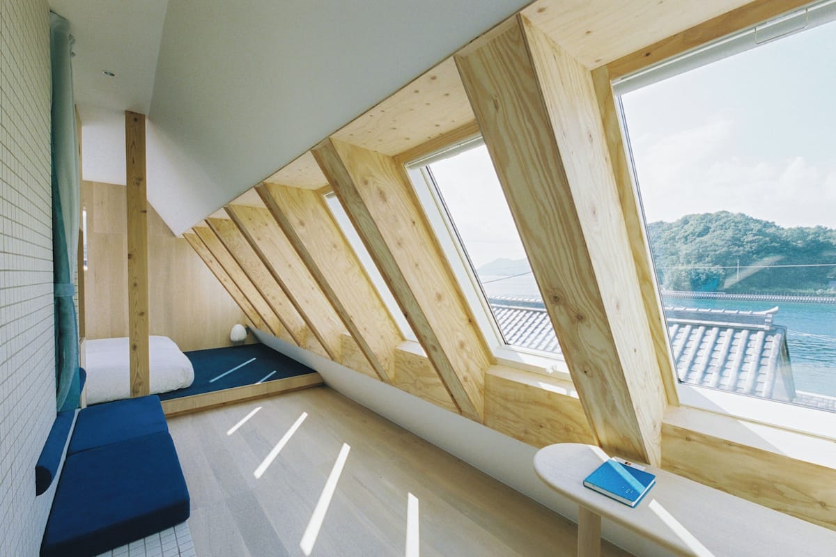 A room with a sloped ceiling, wooden beams, a view of a body of water, and a small table with a book.