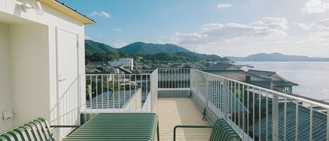 A balcony with a view of a body of water and mountains, green chairs, and a white building.