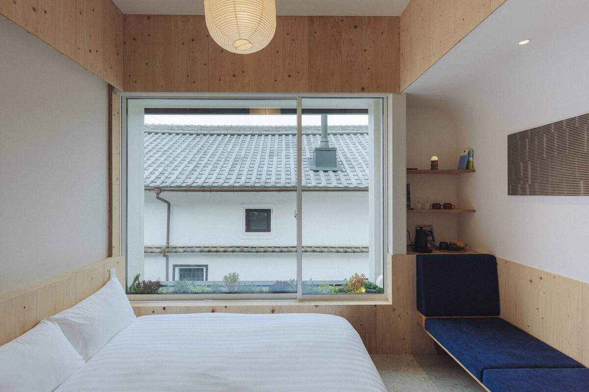 A bedroom with a large window, wooden walls, a bed, a bench, and a shelf with items.