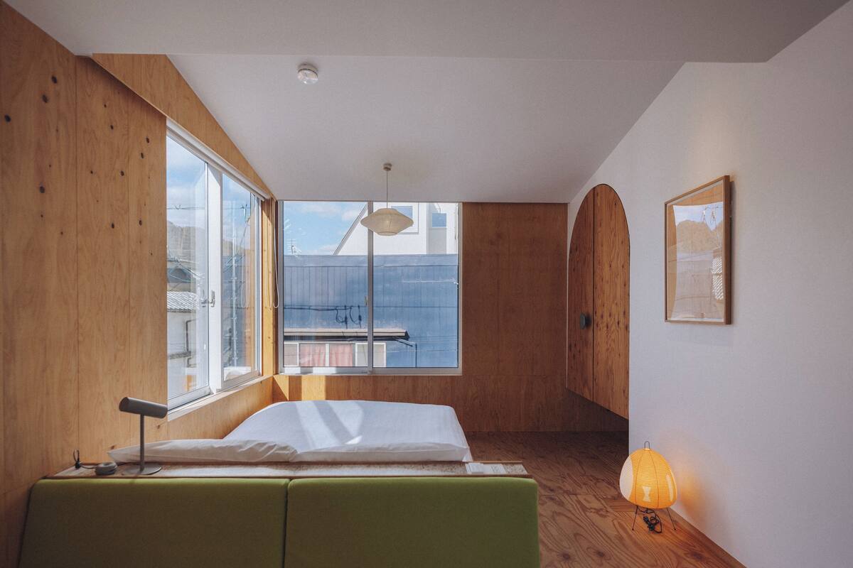 A bedroom with a large window, wooden walls, a bed, a bedside table, and a hanging light fixture.