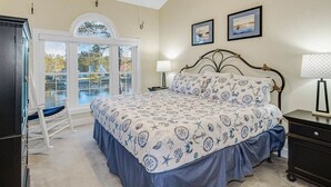 A bedroom with a bed, a nightstand, a lamp, a ceiling fan, and a view through a window. - Cozy two Bedroom Apartment at Riverwalk At Arrowhead (Myrtle Beach)