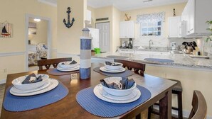 A dining area with a wooden table set for four, a kitchen with white cabinets, and a nautical-themed decor. - Cozy two Bedroom Apartment at Riverwalk At Arrowhead (Myrtle Beach)