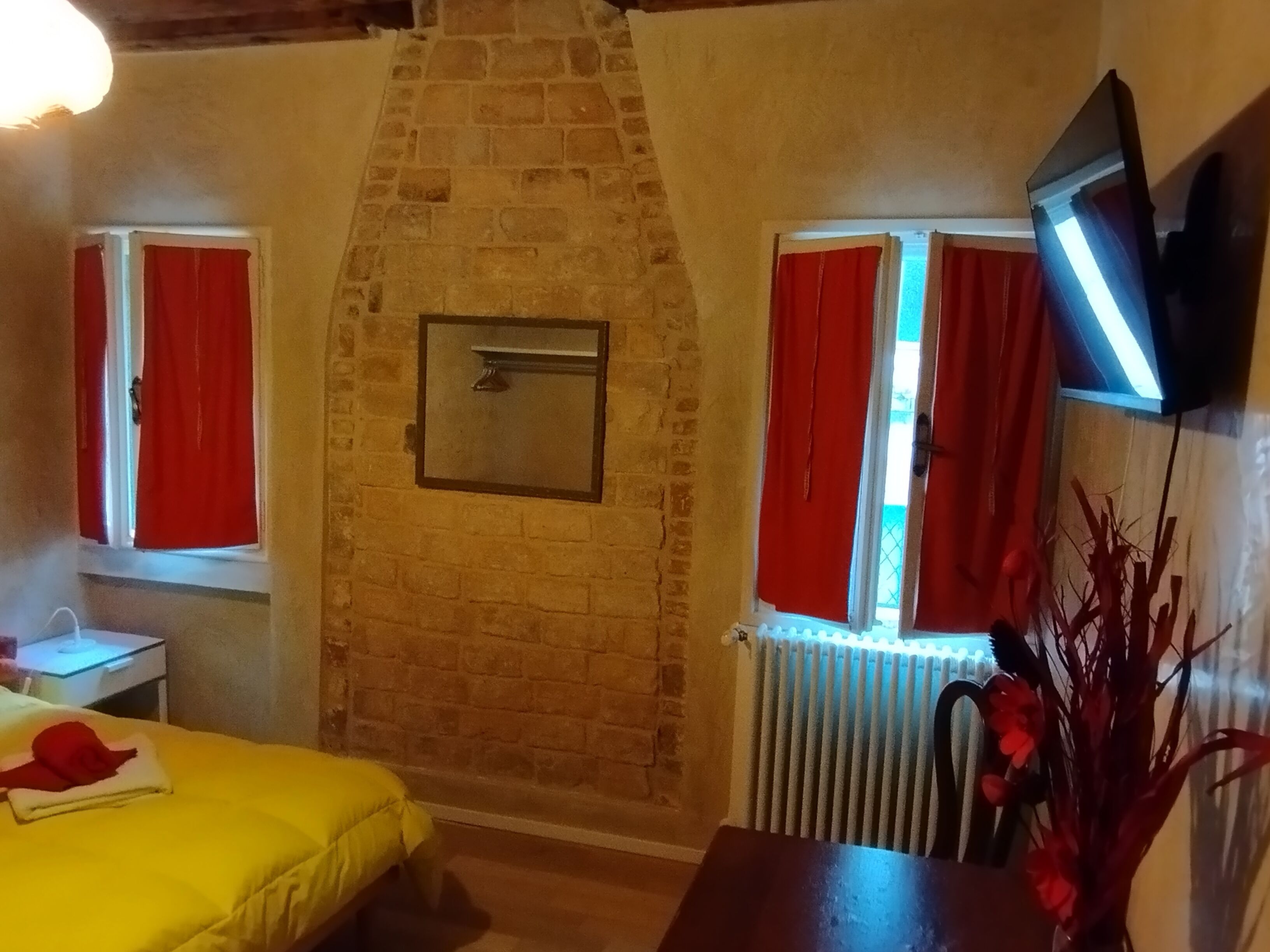 Ein Zimmer mit Kamin aus Ziegelsteinen, roten Vorhängen, einem Bett mit gelber Decke, einem kleinen Tisch und einem an der Wand befestigten Fernseher.