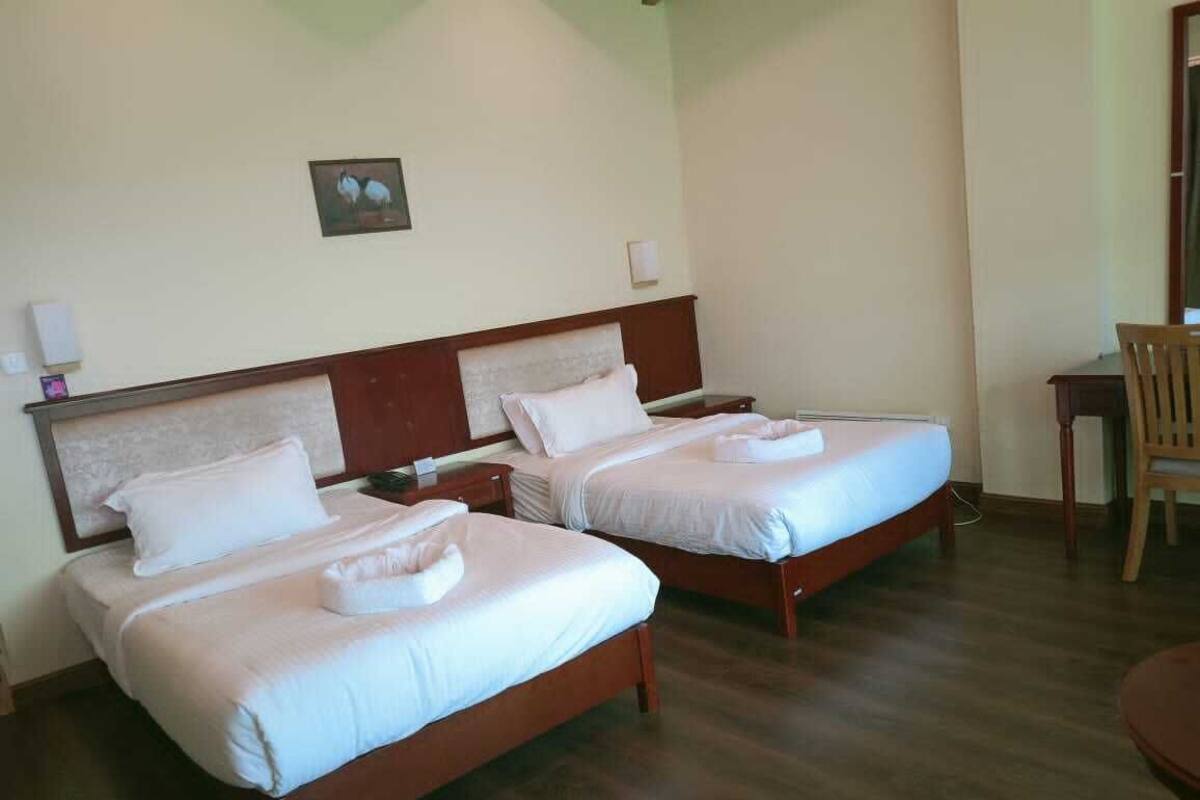 Two single beds with wooden frames and white bedding, a small table, and a chair in a room with wooden flooring and a light-colored wall.