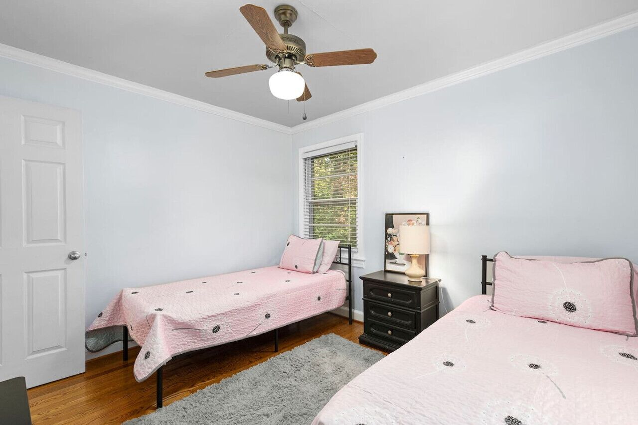 A bedroom with two single beds, a ceiling fan, a window with blinds, a nightstand with a lamp, and a door.