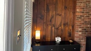 Une pièce avec un mur lambrissé en bois, une commode noire à tiroirs, une petite table avec une lampe et une cheminée en briques.