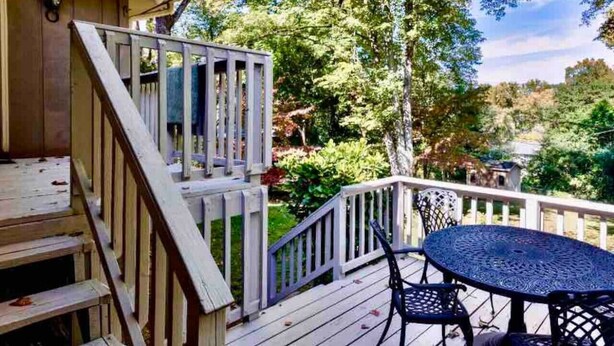 Une terrasse en bois avec un escalier menant à une zone abritée, une table ronde avec des chaises, et une vue sur des arbres et une maison au loin.