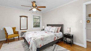 Une chambre à coucher comprenant un lit, une chaise, une table de chevet, un ventilateur de plafond, une fenêtre et un miroir.