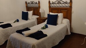 Une chambre d’hôtel avec deux lits simples, chacun agrémenté d’un oreiller bleu et d’une couvre-lit blanche. On y trouve une petite table de chevet avec une lampe et un vase contenant une seule rose rouge. Une table blanche avec un vase contenant une seule rose bleue est également visible.