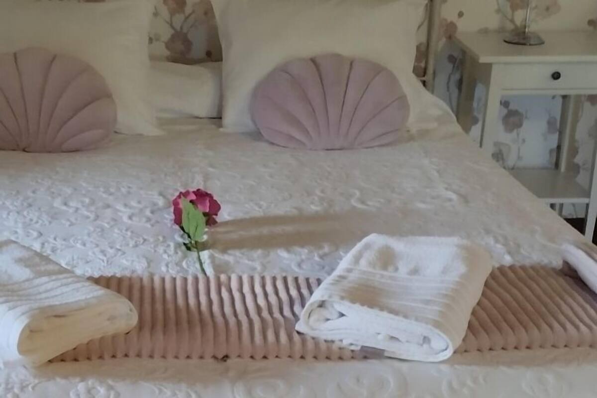 Cama arrumada com uma rosa rosa, uma toalha branca e uma toalha dobrada, com papel de parede floral ao fundo.