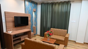 A living room with a flat-screen TV, wooden entertainment unit, brown leather sofa, and a small table with a vase of flowers. - Hotel Olive Crown (Lucknow)