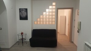 Un salon avec un canapé noir, une table en verre sur laquelle se trouve une bouteille, un tableau encadré représentant un lion et un escalier menant à une autre pièce.