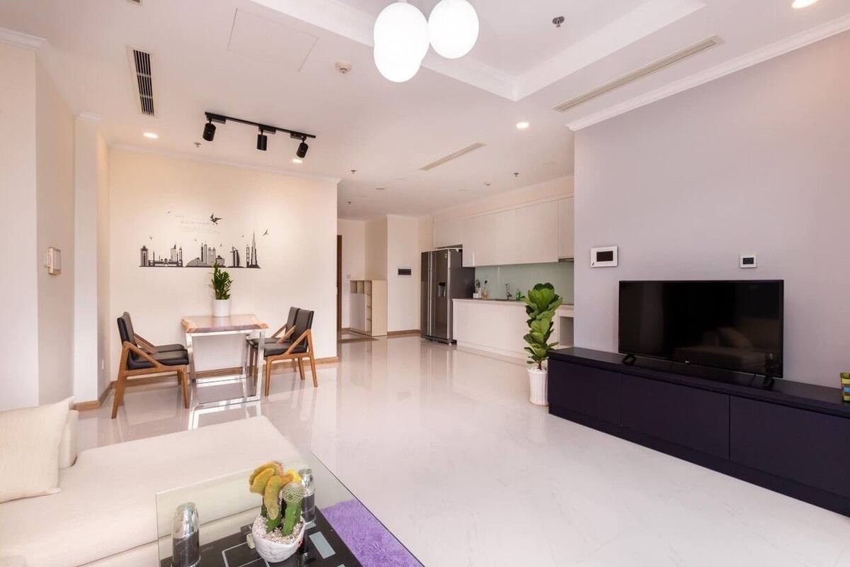 A modern living room with a sofa, a coffee table with a vase, a dining area with chairs and a table, a kitchen with a refrigerator, and a TV mounted on the wall.