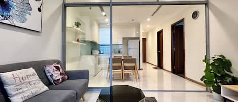 Un salón moderno con un sofá gris, una mesa de centro negra y una puerta de vidrio que da a una cocina y zona de comedor.