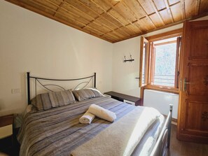 A bedroom with a wooden ceiling, a bed with a striped bedspread, a rolled towel, a nightstand, a small table, a wooden door, and a window with a view of trees. - Grand Hotel Ceresole Reale Bill Apartment (Ceresole Reale)
