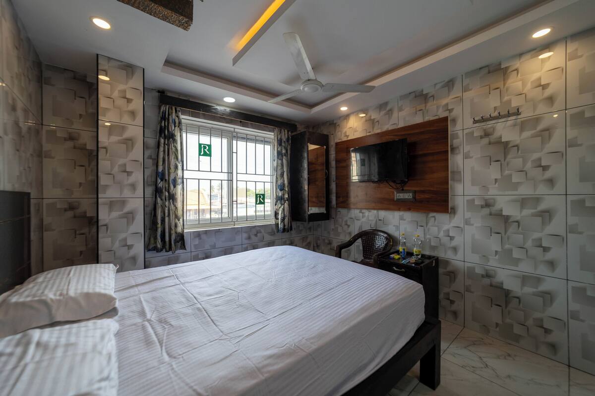 A modern bedroom with a large bed, a ceiling fan, a TV mounted on the wall, and a window with curtains.
