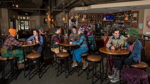 Eine Bar mit runden Tischen und Hockern, Holzregalen mit Flaschen und Pendelleuchten.