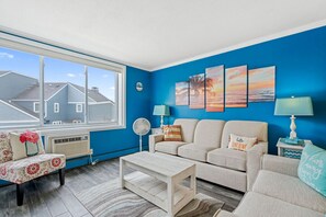 A living room with a large window, blue walls, a beige sofa set, a small table, and a fan.
