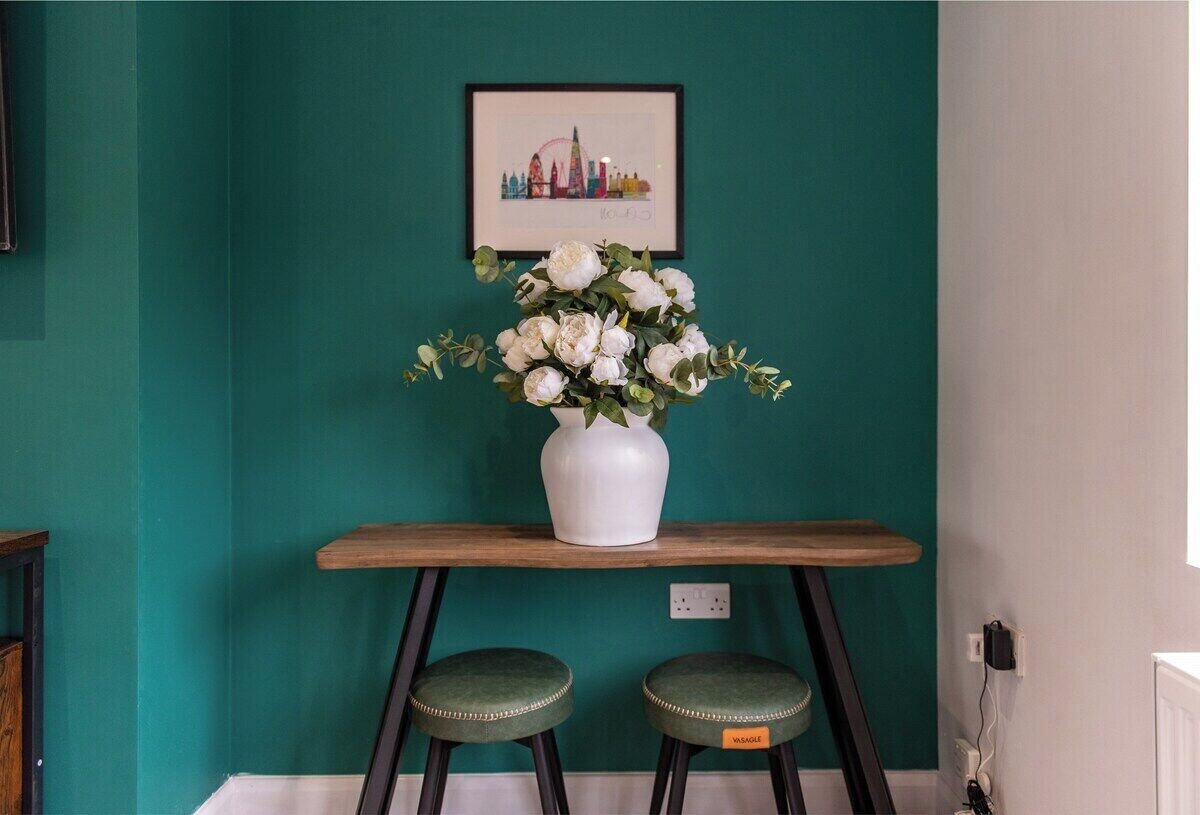 A teal wall with a framed cityscape artwork, a white vase with flowers, and two green stools.