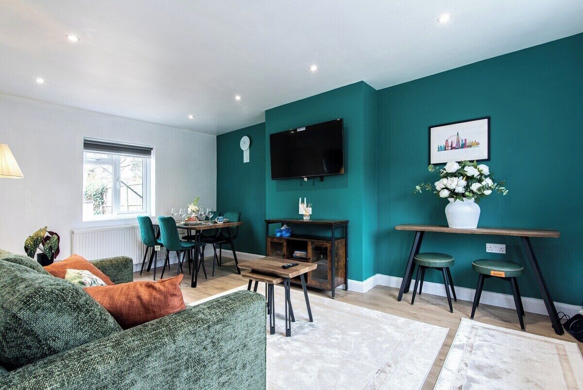 A living room with teal walls, a flat-screen TV, a dining area with chairs, and a sofa with cushions.