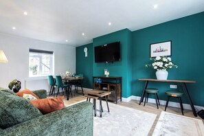 A living room with teal walls, a flat-screen TV, a dining area with chairs, and a sofa with cushions.