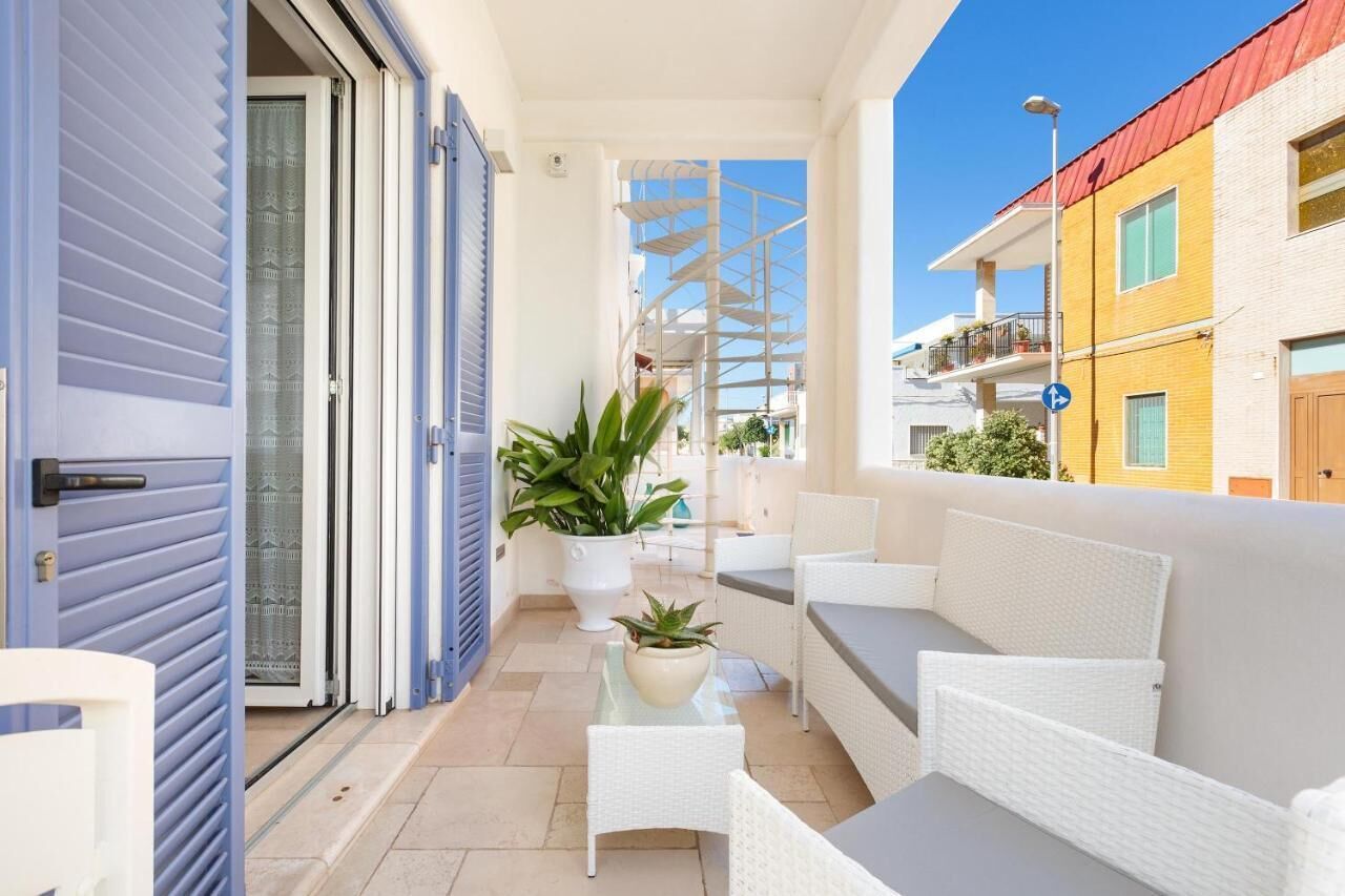 Un balcón con mobiliario de mimbre blanco, una planta en maceta y vistas a edificios vecinos.