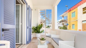 Un balcón con mobiliario de mimbre blanco, una planta en maceta y vistas a edificios vecinos.