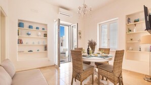 Un comedor con sillas de mimbre, una mesa redonda y una araña. Se observan estanterías con libros y objetos decorativos en las paredes.