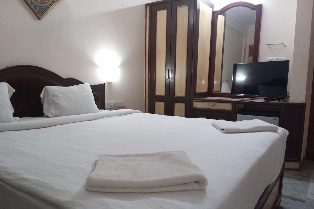 A neatly made bed with a folded towel, a wooden wardrobe, a television on a wooden stand, and a wall-mounted light.