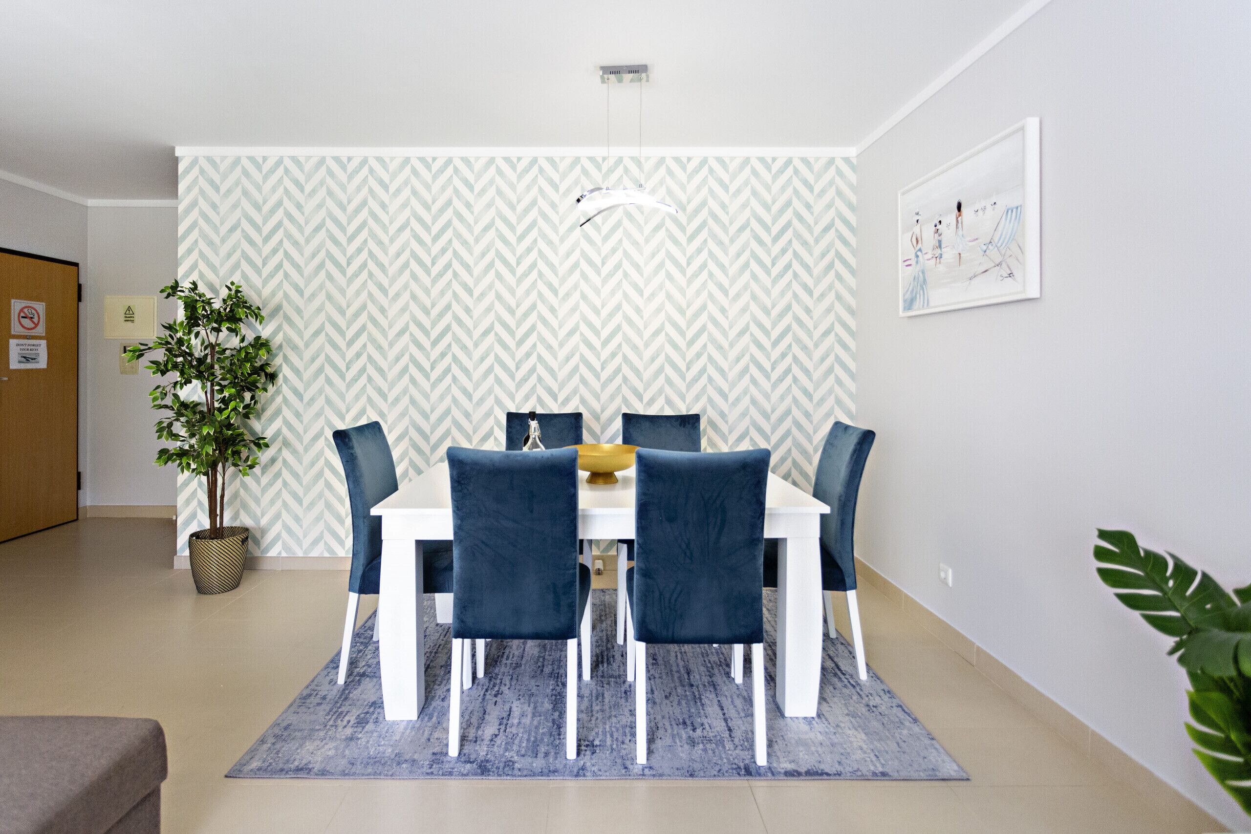 Un coin repas avec une table blanche et des chaises bleues, un papier peint à motifs et une plante en pot.