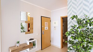 Un couloir avec un banc en bois, une petite étagère avec des plantes, une porte sur laquelle est apposé un panneau « interdiction de fumer » et une plante en pot.