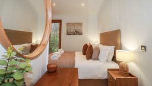 A modern bedroom with a large mirror, a wooden headboard, a bedside table with a lamp, and a view of a garden through a window. - 1 Streamside (Looe)