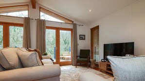 A spacious living room with a sofa, a television, and a view through large glass doors to an outdoor area. - 1 Streamside (Looe)