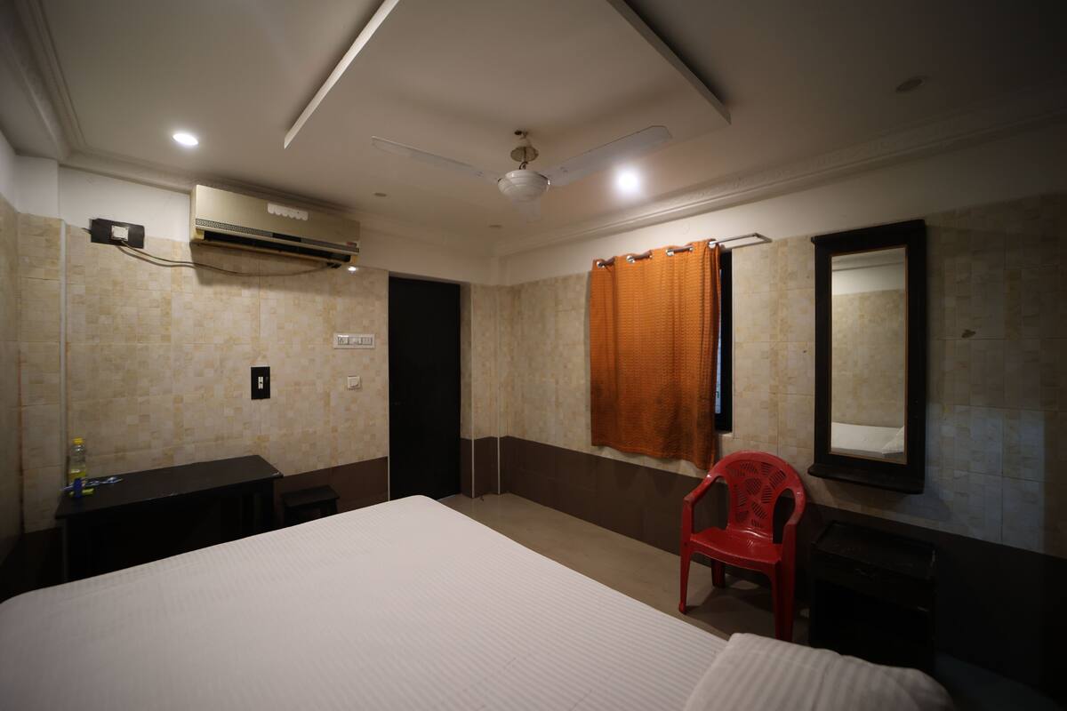 A hotel room with a bed, a desk, a chair, an air conditioning unit, a mirror, and a shower curtain.