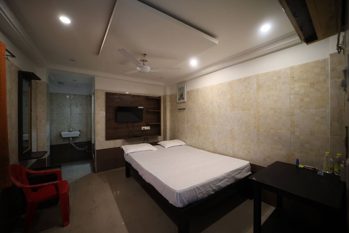 A hotel room with a bed, a desk, a chair, a TV, and a bathroom with a sink and mirror.